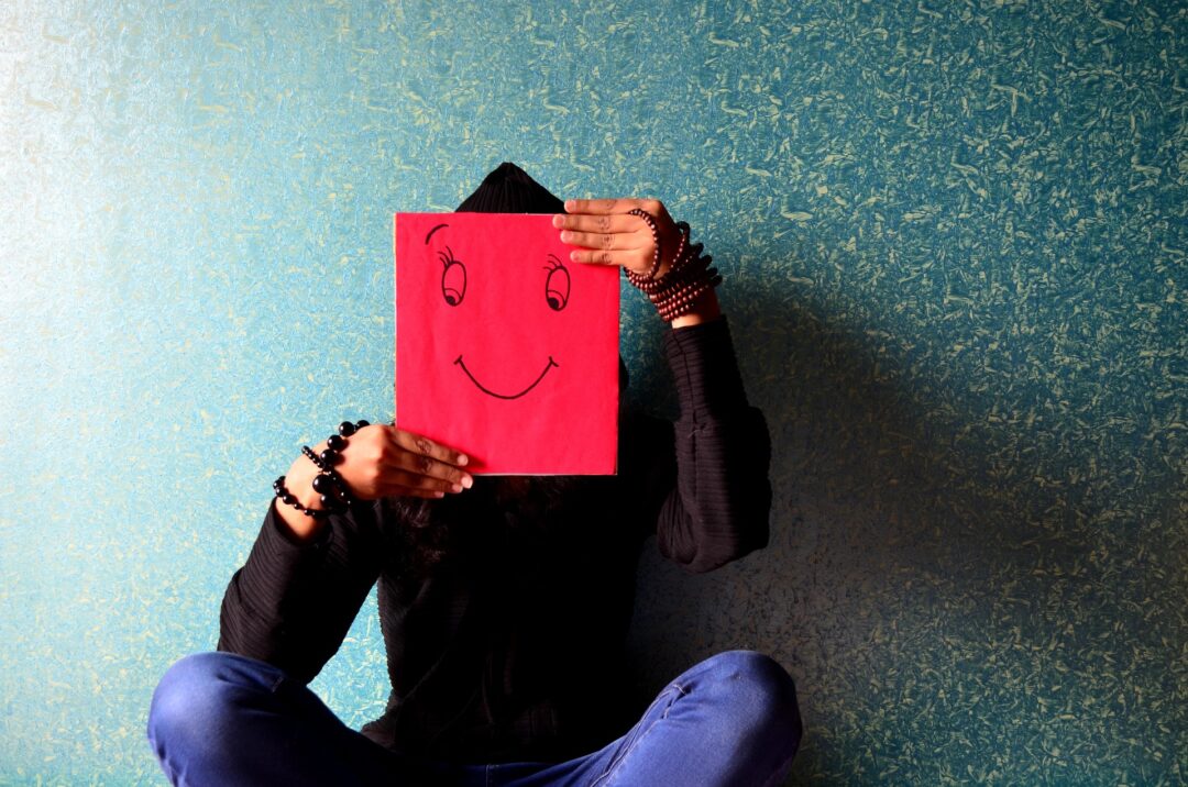 An image of a person covering their face by a smiley face drawn on a red sheet of paper